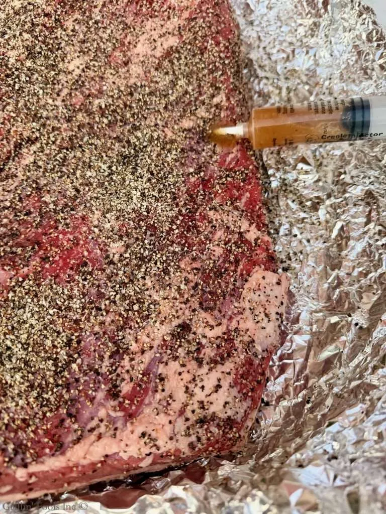 Beef brisket crusted with SPG seasoning on aluminum foil with a marinade syringe nearby, ready to cook.