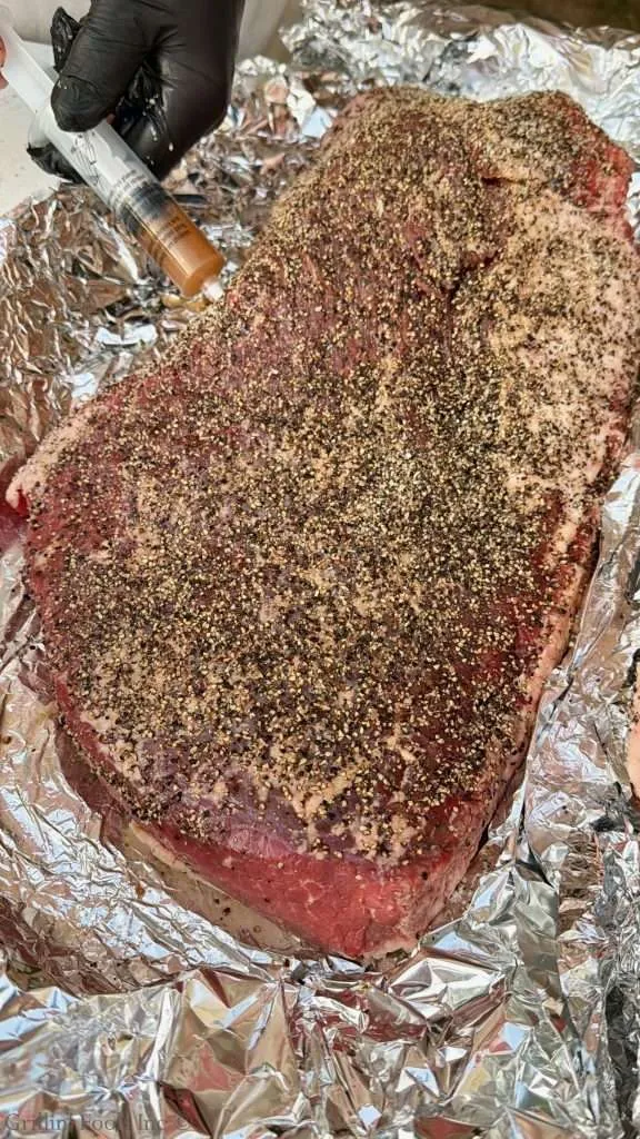 Beef brisket covered in cracked pepper sits on aluminum foil as a gloved hand injects marinade with a syringe.