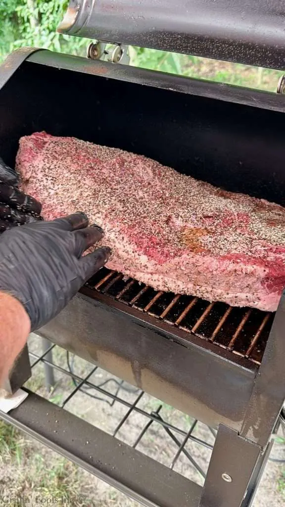 Hands wearing black gloves handling a large seasoned brisket on a smoker grill outdoors.