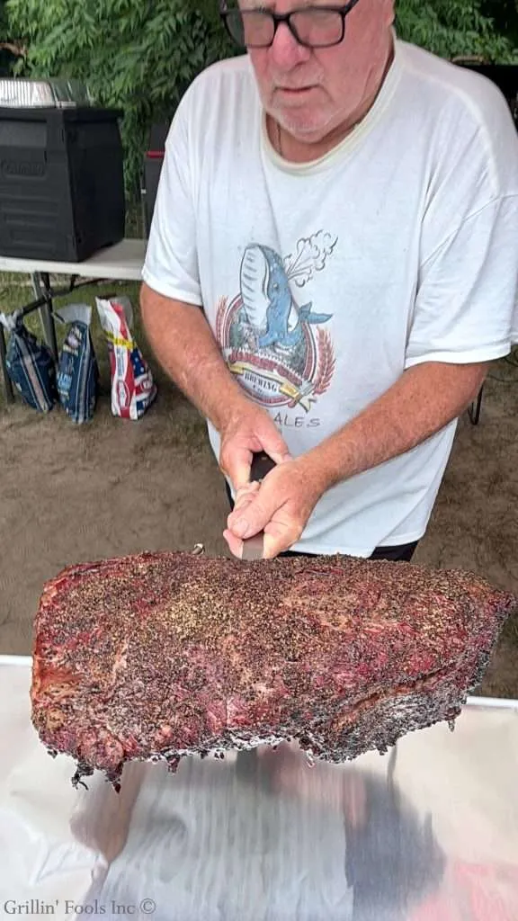 Older man wearing glasses carrying a large smoked brisket on a spatula to a table outdoors, covered in foil