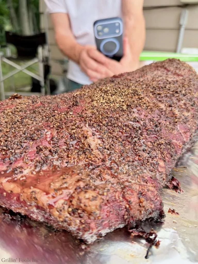 Close-up of pepper-crusted smoked brisket resting on a tray with a blurred person holding a smartphone in the background.