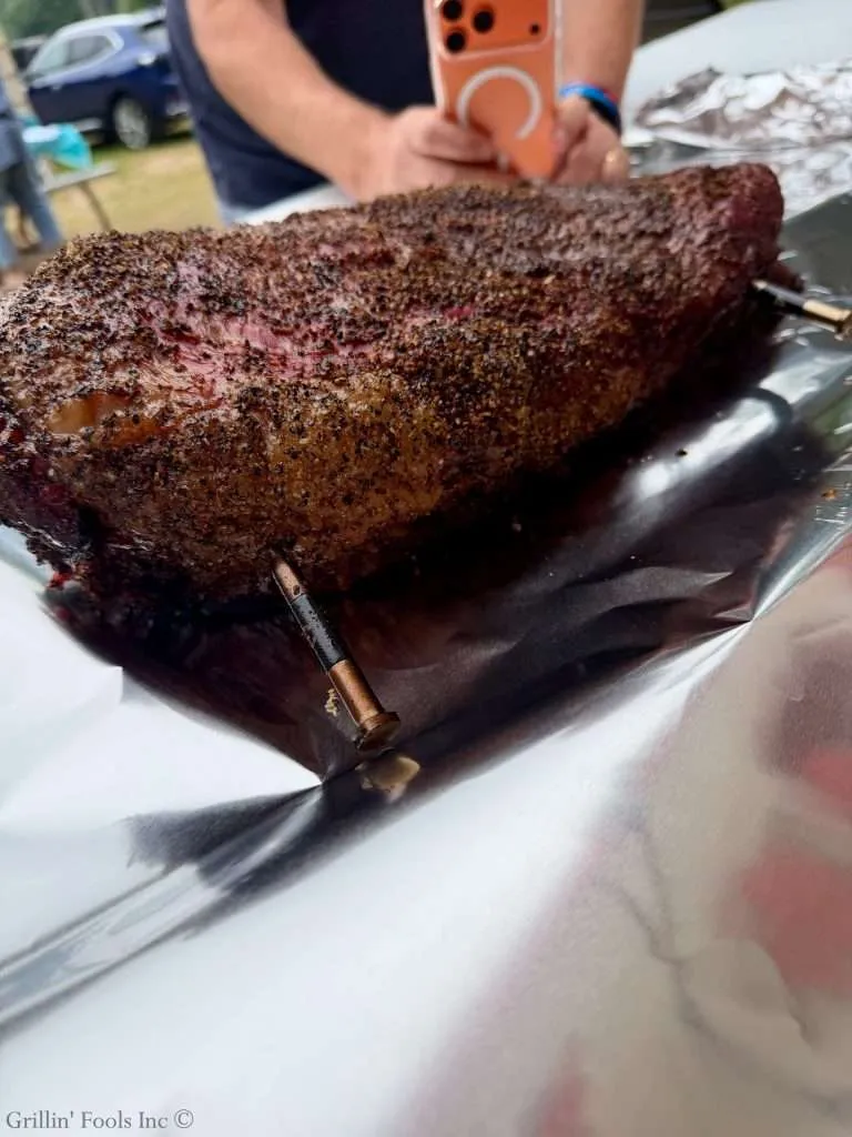 Smoked, pepper-crusted brisket resting on aluminum foil, with a person holding an orange smartphone in the background.