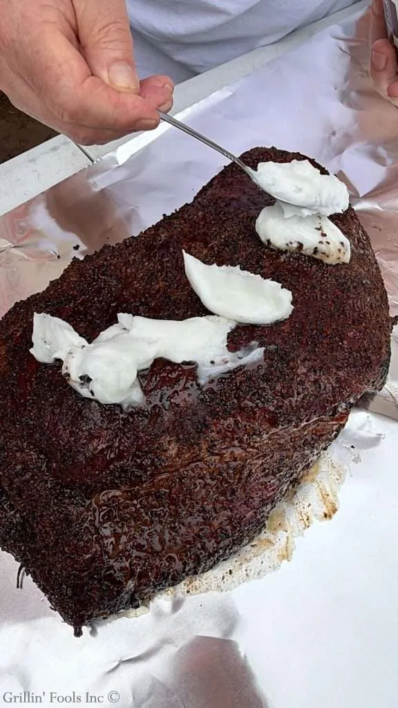 Close-up of a smoked beef brisket with a dark bark, having dollops of beef tallow added to the top