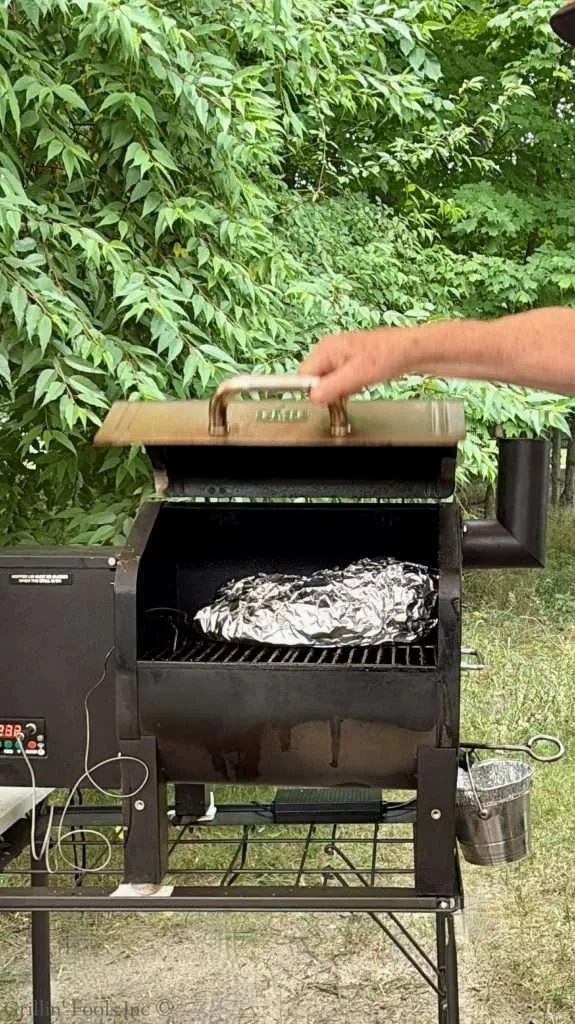 Outdoor charcoal grill with the lid open; a hand is lifting the lid and a foil-wrapped package rests on the grate inside. Lush green bushes in the background.