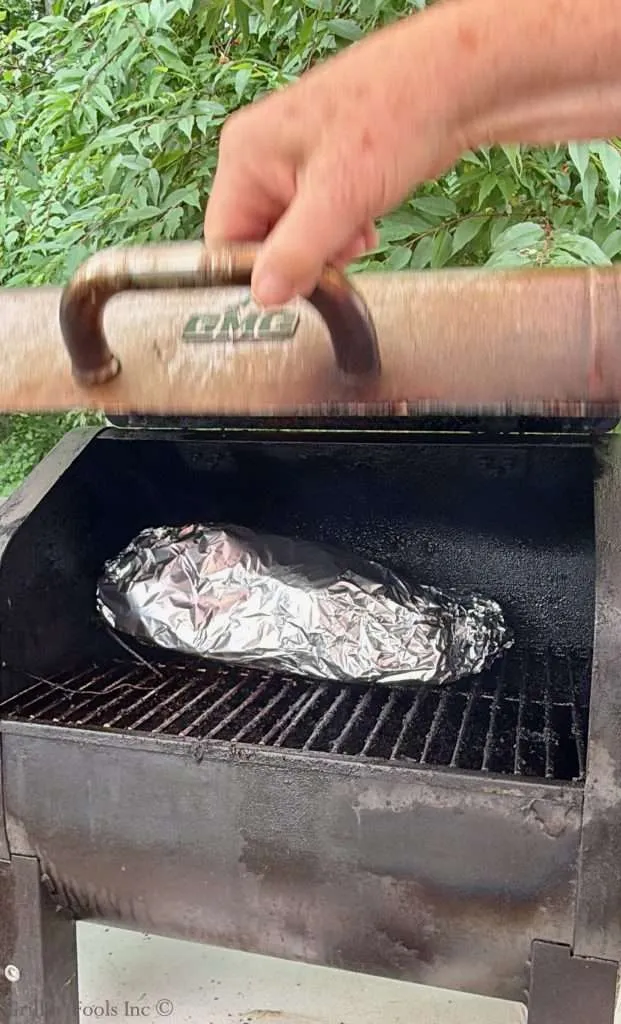Hand lifting a copper barbecue lid to reveal a foil-wrapped item inside, with greenery in the background.