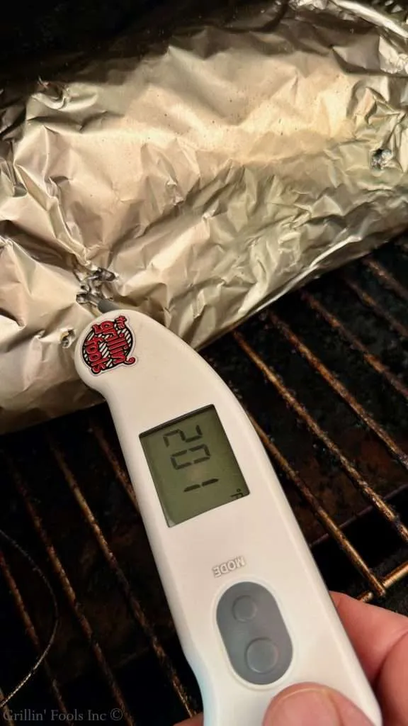 Digital meat thermometer resting on a grill next to crumpled aluminum foil-wrapped food, showing a 201F on the screen.