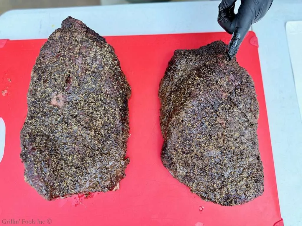 Two smoked beef briskets with a dark peppery crust sit on a red cutting board; a gloved hand points at the one on the right