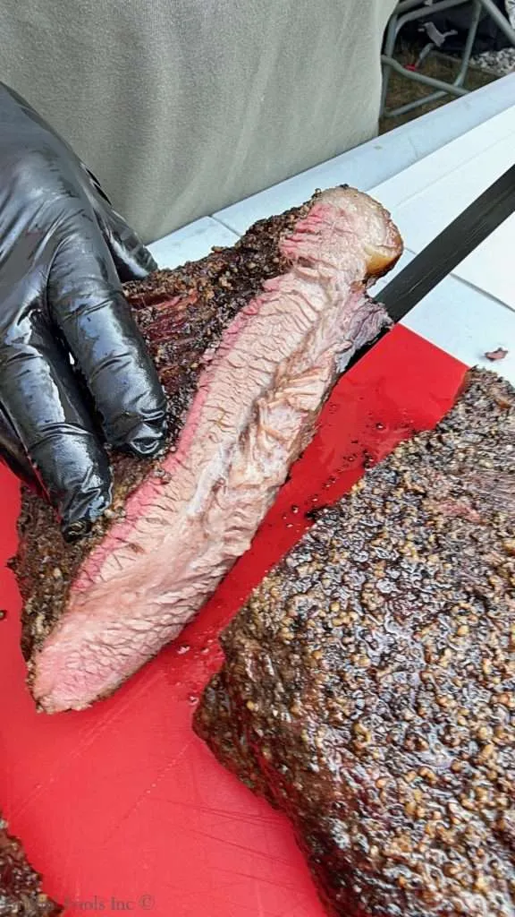 Slices of smoked beef brisket with a pink smoke ring and dark crust, held by a gloved hand on a red cutting board.