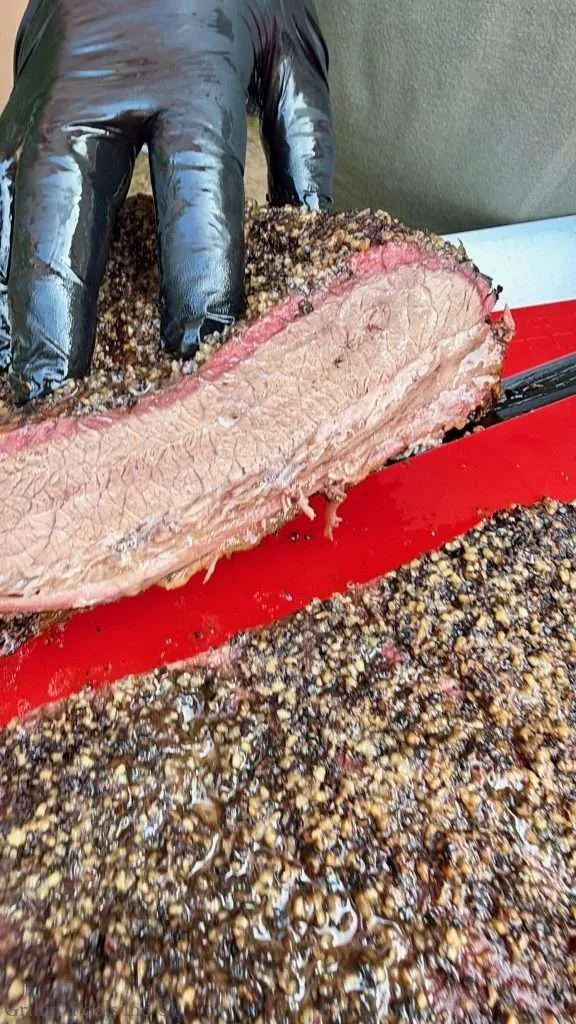 Sliced smoked beef brisket with a pepper crust and pink smoke ring, being cut with gloved hands on a red cutting board.