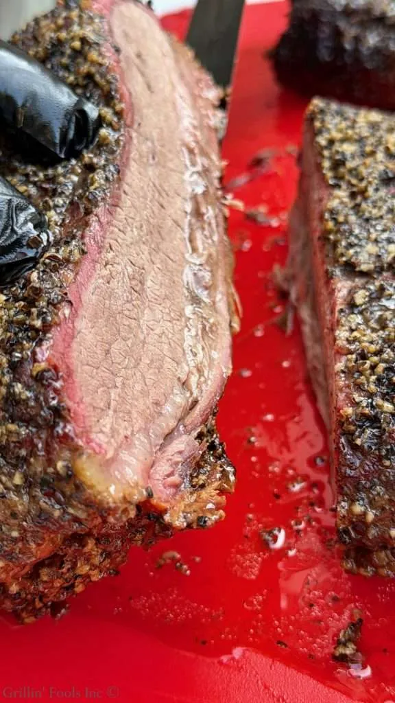 Sliced pepper-crusted smoked beef brisket on a red cutting board, showing a pink smoke ring and juicy interior.