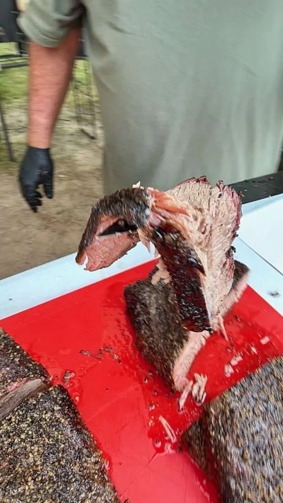 Sliced smoked beef brisket with a dark crust on a red cutting board; a gloved worker stands in the background.