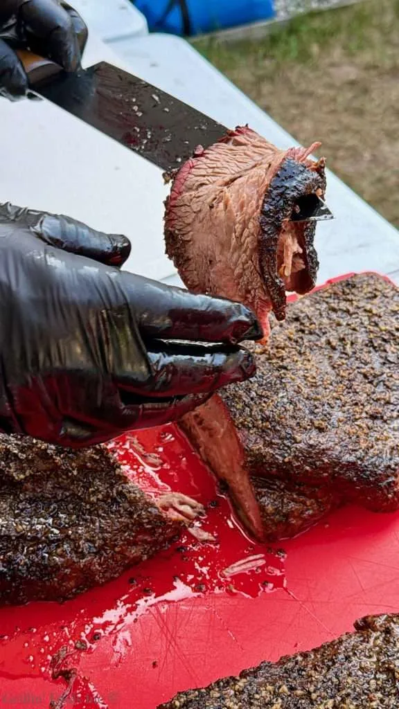 Close-up of gloved hands carving a large smoked roast with a knife, exposing a pink interior and brown crust on a red cutting board.