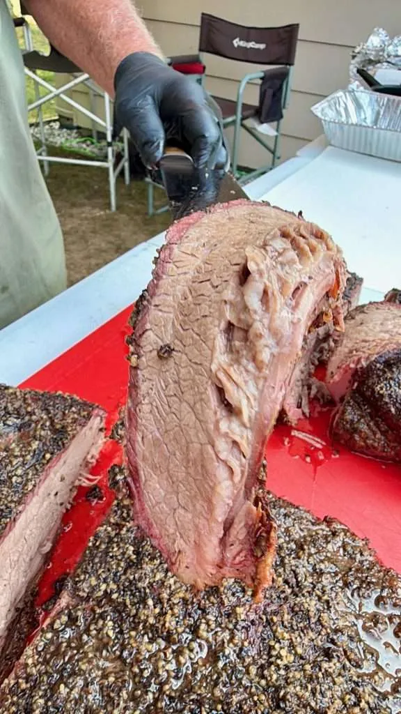 Smoked beef brisket being sliced on a red cutting board outside, a gloved hand holds a knife to carve the meat.