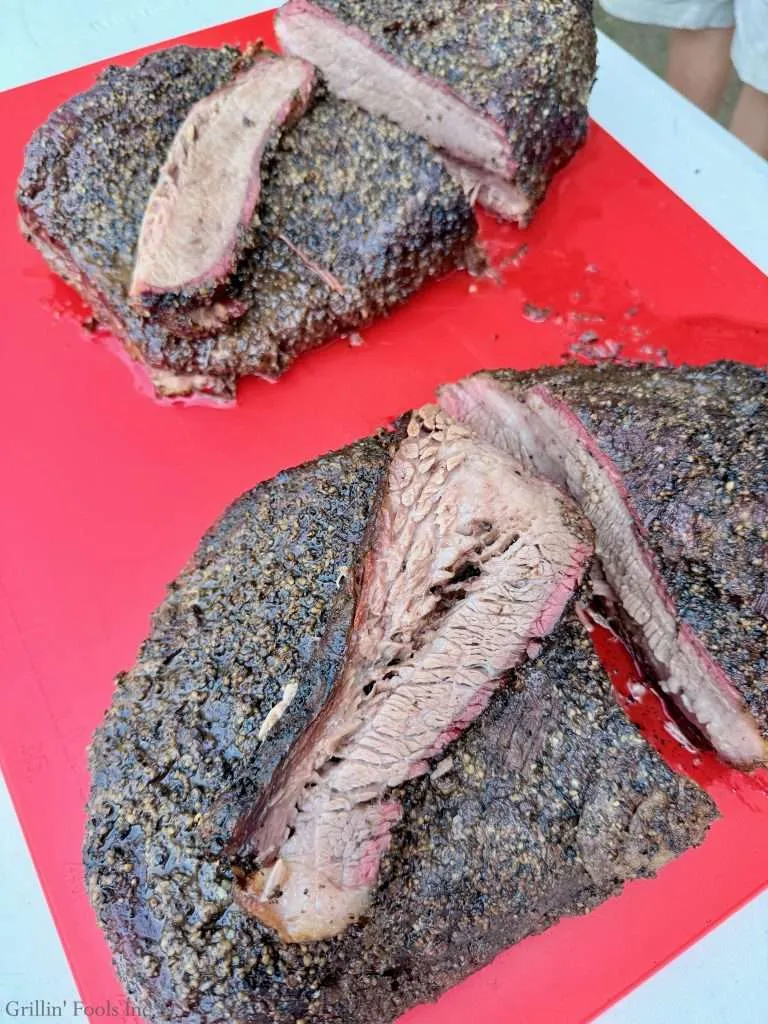 Sliced smoked beef briskets with a dark pepper crust and pink smoke-ring interior on a red cutting board, the one closer to the camera got the dry brine overnight