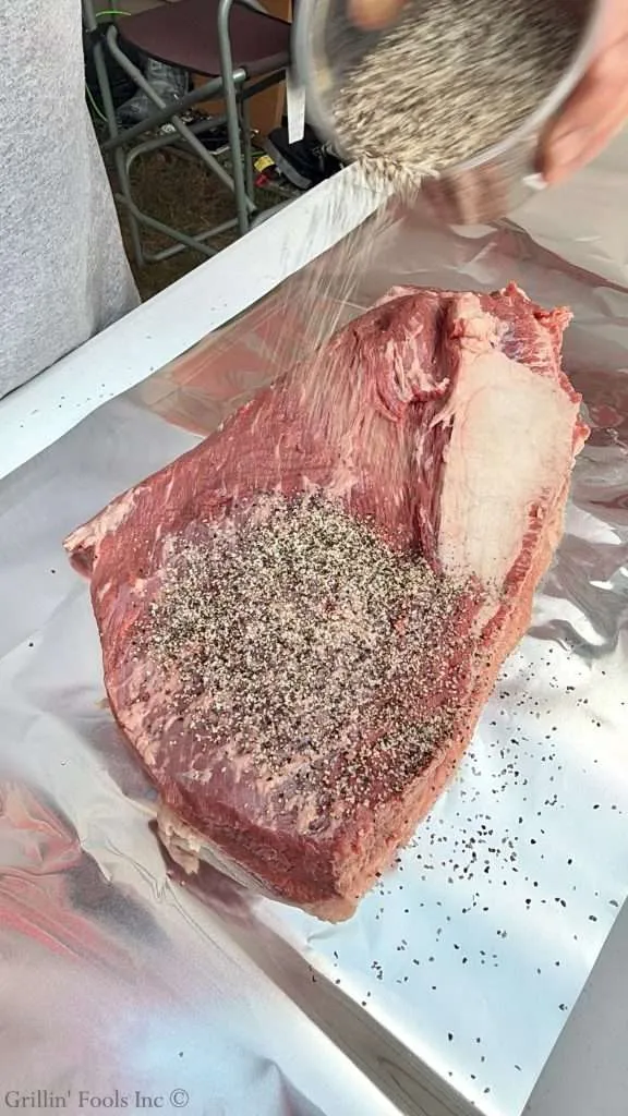 Seasoning a raw packer brisket with SPG seasoning from a plastic jaw, all on a sheet of aluminum foil that will have the dry brine overnight