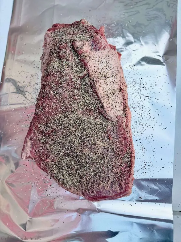 A raw, whole packer beef brisket seasoned with an SPG rub, all on a sheet of aluminum foil taken from above