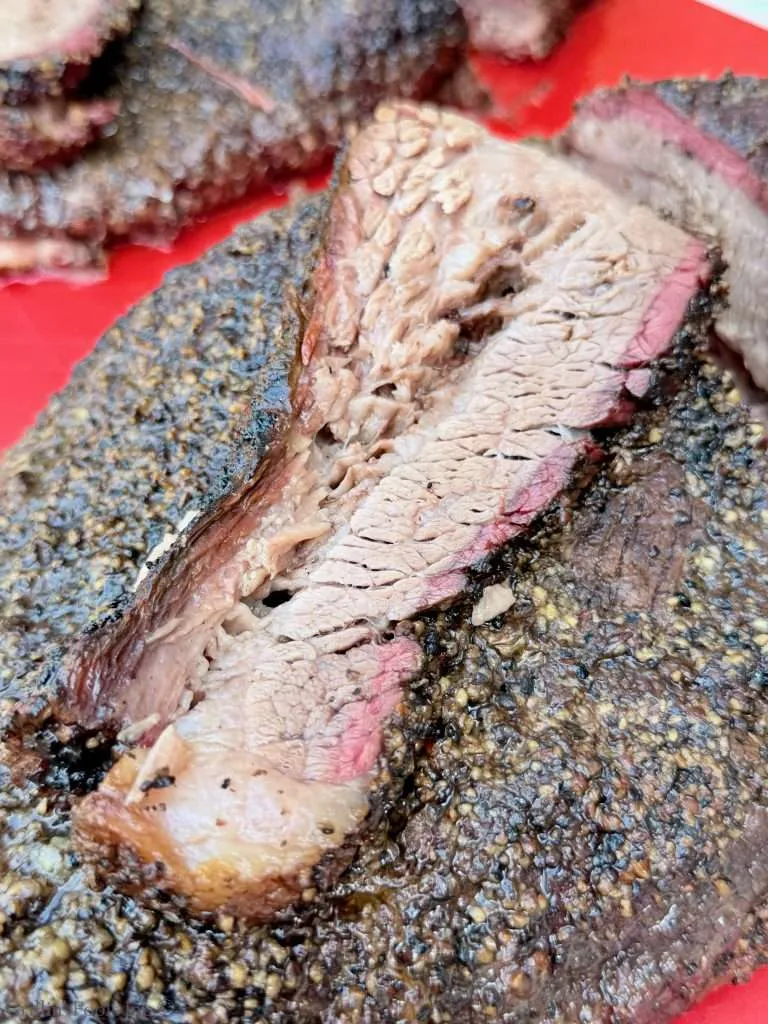 Close-up of smoked beef brisket slices with a dark pepper crust, pink smoke ring, and juicy interior on a red plate. This brisket got the dry brine overnight
