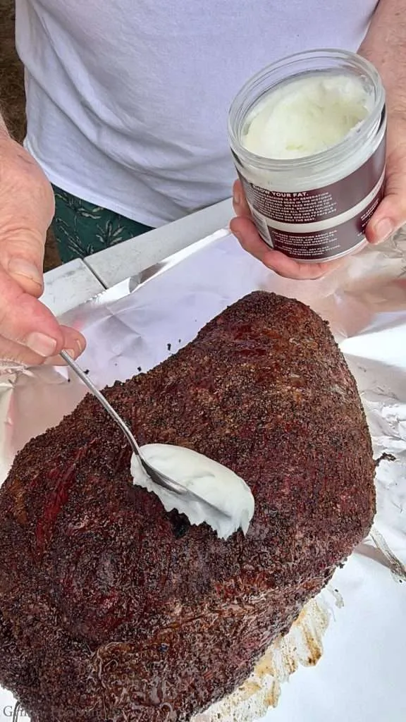 Person applying beef tallow onto a large, dark crusted smoked brisket using a spoon and jar nearby