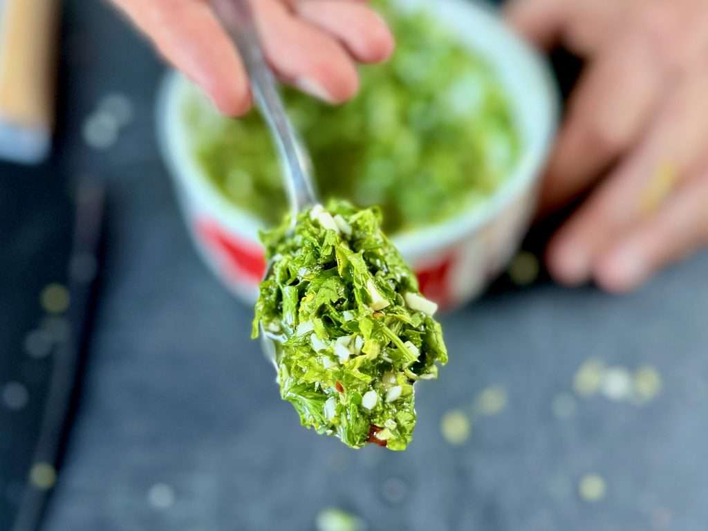 A spoonful of chimichurri sauce held over a blurry bowl of chimichurri