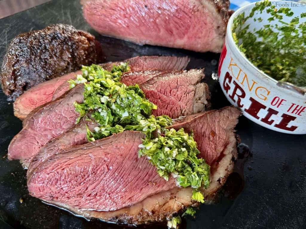 Sliced Picanha Roast with Chimichurri spooned over the top, with a bowl that reads "King of the Grill" holding more chimi next to the beef