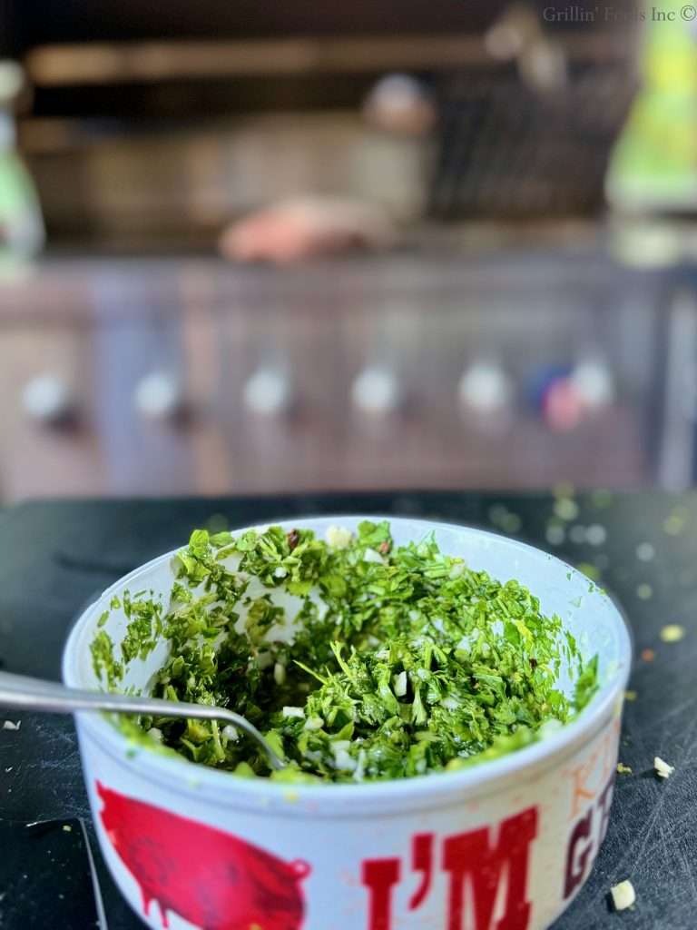 A bowl of easy chimichurri sauce in front of a blurred, stainless steel grill with a large roast on top