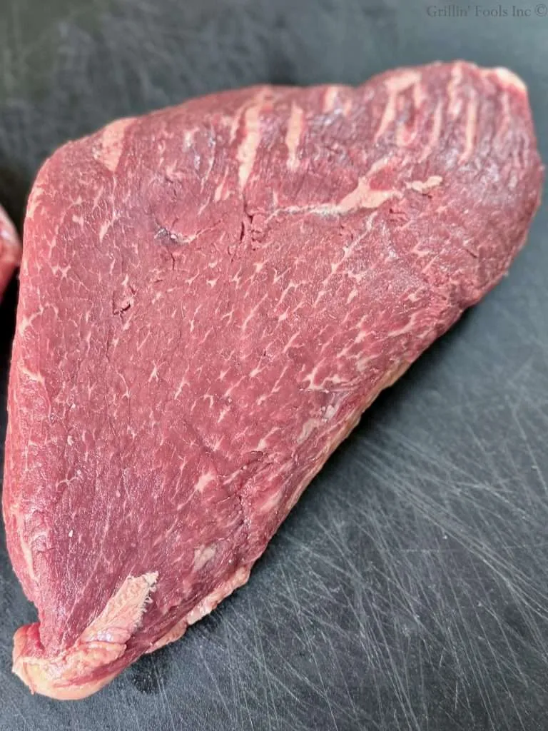 A raw picanha roast, shown fat cap down, meat side up, on a black cutting board from above.