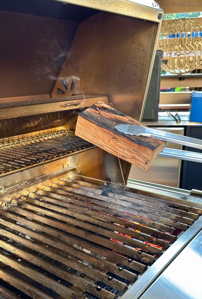 A chunk of smoke wood smoldering, while being held by a pair of tongs over some hot coals in a charcoal grill