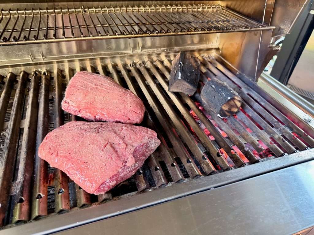 Two raw, seasoned picanha roasts cooking indirect, near some hot coals and a couple chunks of blackened smoke wood.
