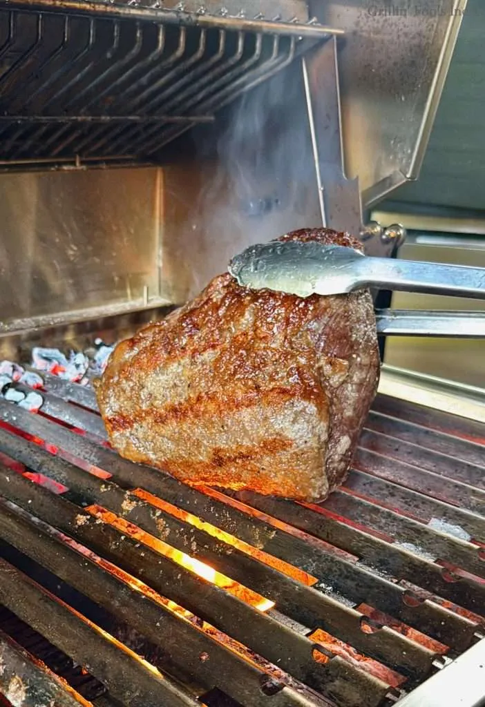 A picanha roast, browning over the coals, being held up by tongs