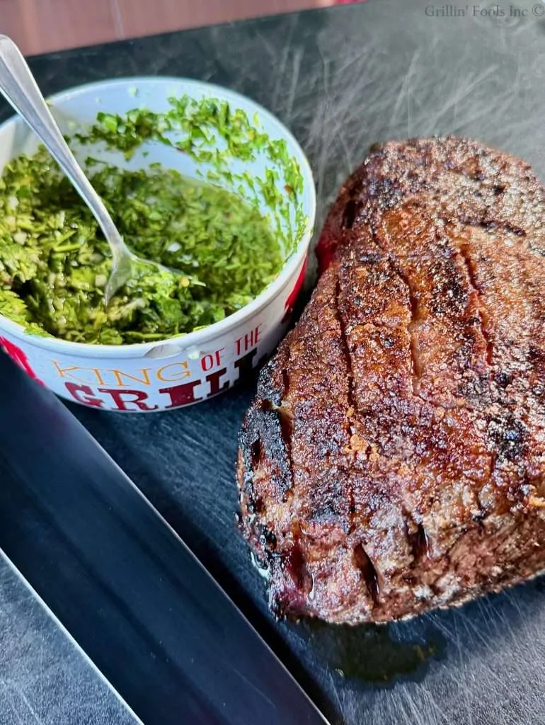 A roasted picaña next to a bowl of chimichurri and a black-bladed knife.