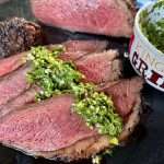 Sliced Picanha Roast with Chimichurri spooned over the top, with a bowl that reads "King of the Grill" holding more chimi next to the beef