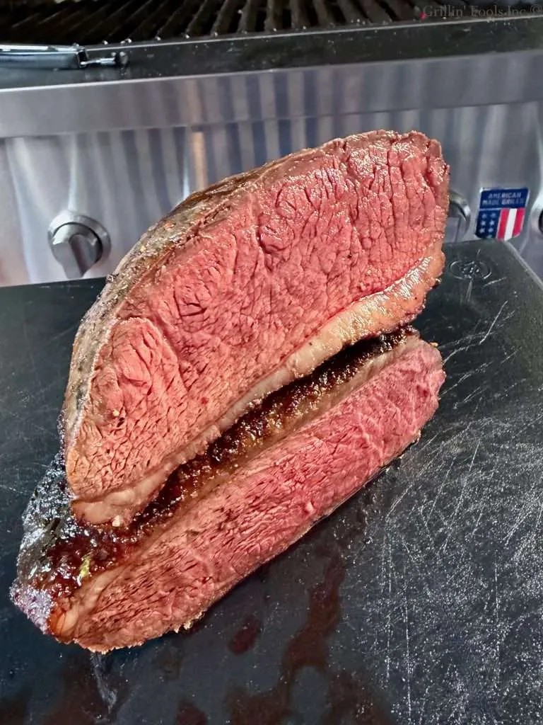 A picaña roast, sliced in half with one half on top of the other, a stainless grill is in the background