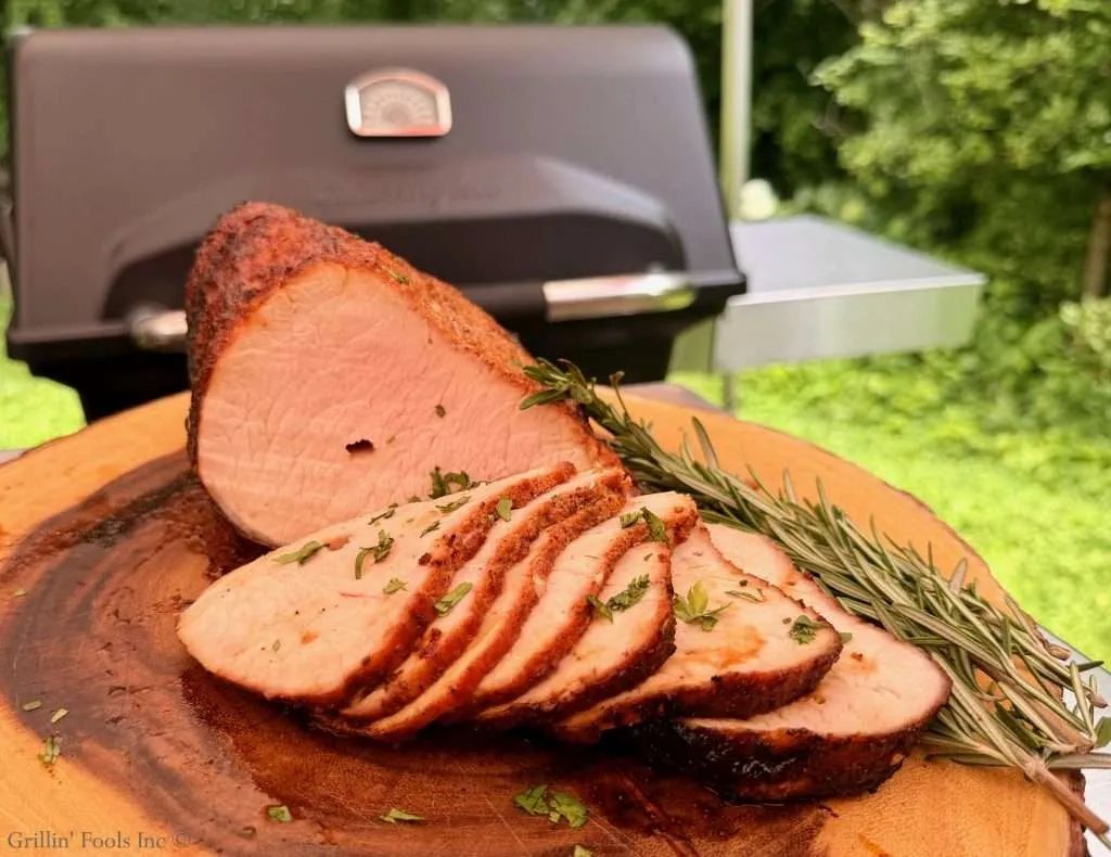 Sliced roasted pork loin arranged on a wooden cutting board with rosemary sprigs, outdoors near a grill in a sunny backyard.