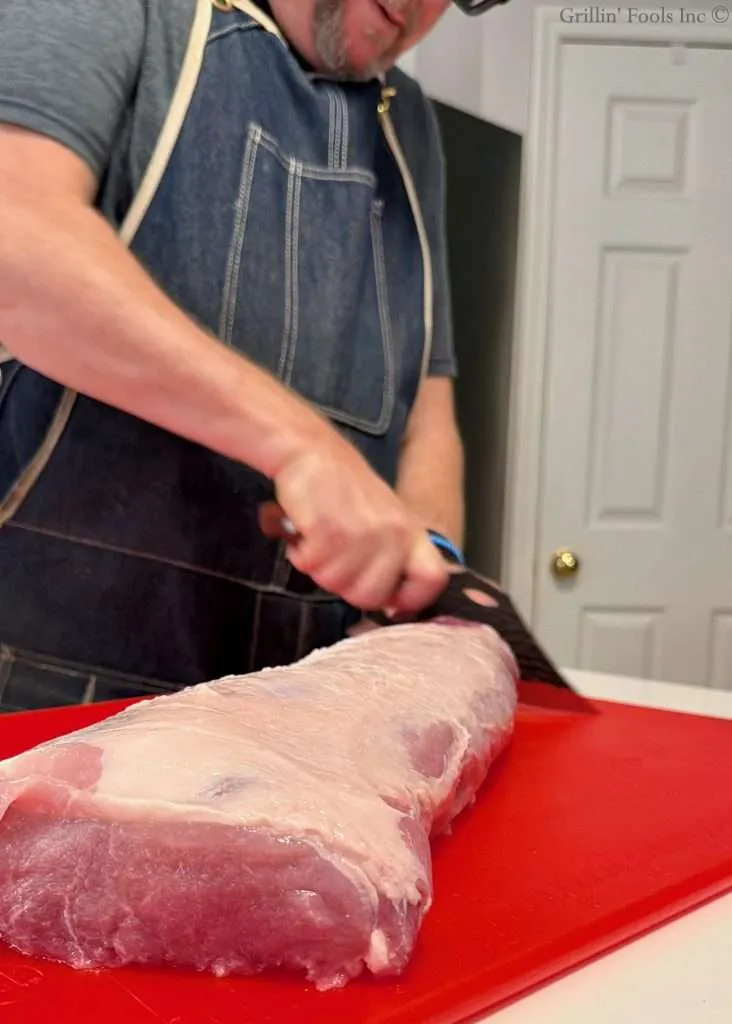 Adult wearing a denim apron slicing a large pork loin cut on a bright red cutting board in a kitchen.