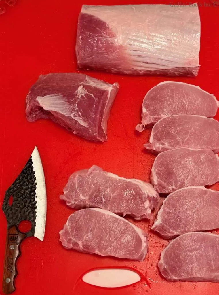 Raw pork chops, and two pork roasts arranged on a red cutting board with a knife nearby, ready for prep.