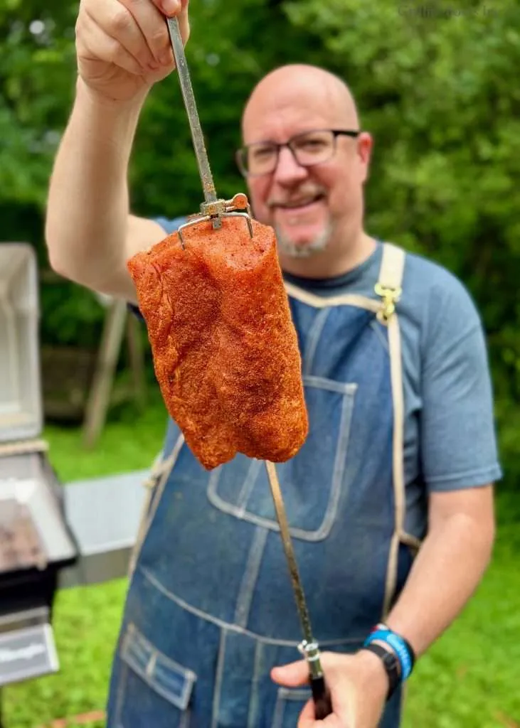 Smiling man in a denim apron outdoors holding a large, seasoned pork loin on a rotisserie spit in front of him.