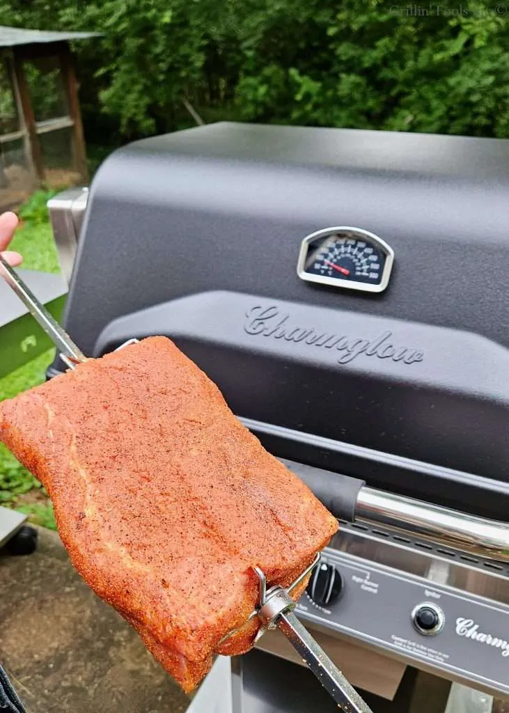 Close-up of a seasoned pork loin on a metal skewer held in front of a Charmglow grill with a built-in thermometer on the hood, outdoors.