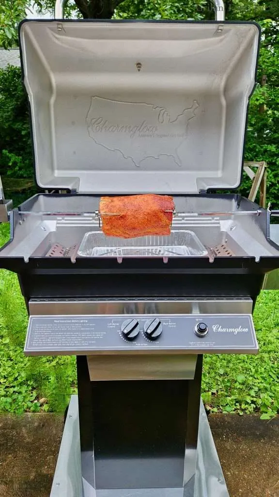 Open-lid Charmglow grill with a rotisserie spit holding a seasoned, but raw, pork roast over a drip pan; stainless control panel visible, outdoor greenery in the background.