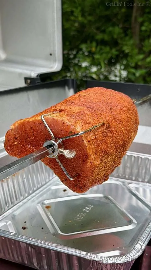 Seasoned roast on a rotisserie spit clamped in a barbecue grill, over an aluminum tray outdoors