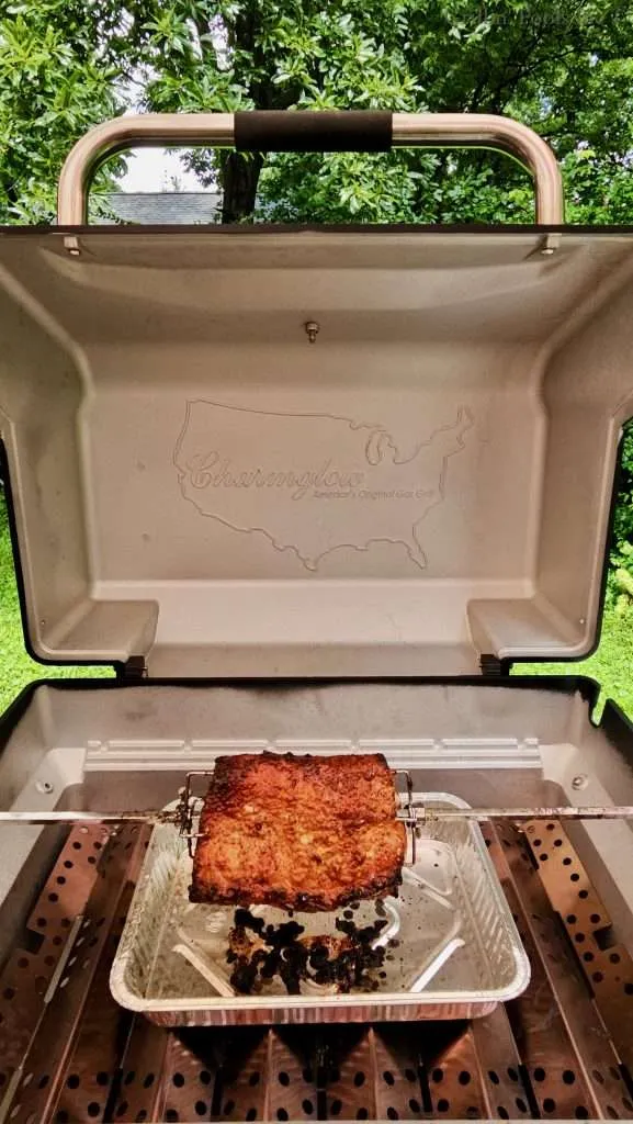 Open gas grill with the lid up, a roasted, brined, rotisserie pork loin on a spit resting in a foil tray, outdoors in a backyard with green trees behind.