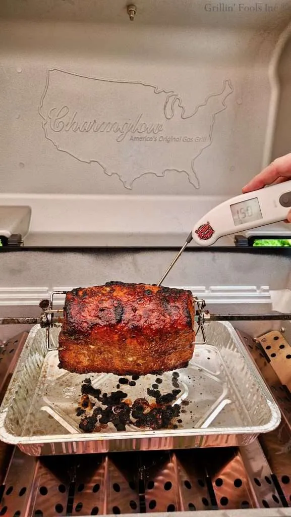 Whole roasted pork loin on a rotisserie inside a grill, with a map of the United States etched on the back wall and a thermometer showing about 150°F nearby.
