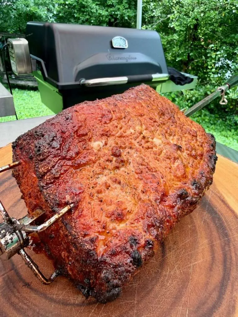 A skewered whole roasted pork loin on a with a golden brown crust in front of a Charmglow gas grill,