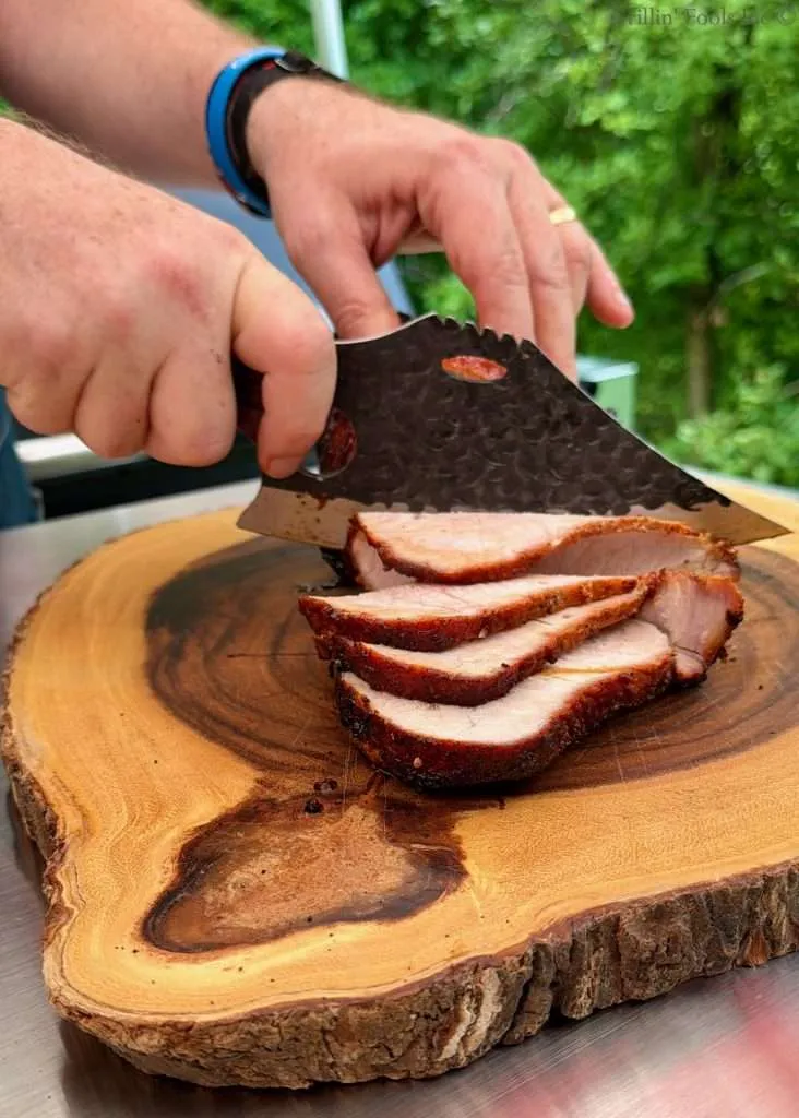 Hands slice a smoked pork loin roast into thin pieces on a round wooden cutting board outdoors, using a wicked looking knife.