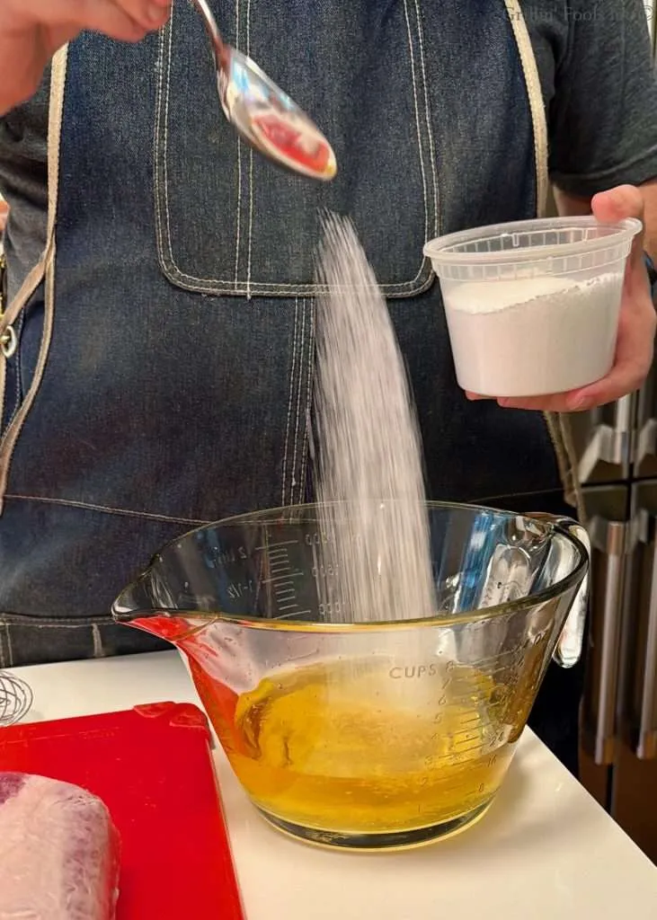 Person in a denim apron pours salt from a spoon into a glass measuring bowl of apple juice on a kitchen counter; a pork loin, fat cap up, on a red cutting board nearby.