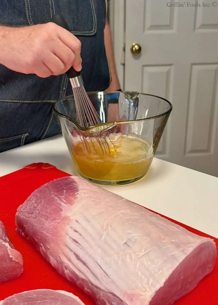 Person whisking apple juice and salt in a glass mixing bowl; raw pork loin on a red cutting board nearby in a kitchen.