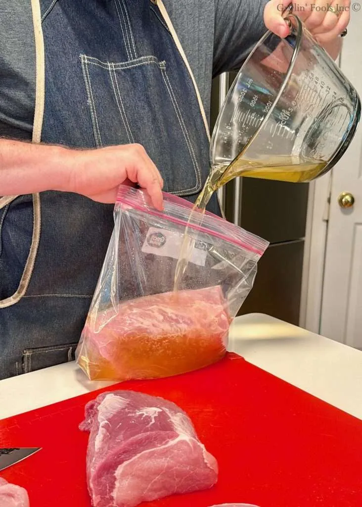Person in a denim apron pours an apple juice brine from a measuring jug into a resealable plastic bag containing a raw pork loin on a red cutting board in a kitchen.