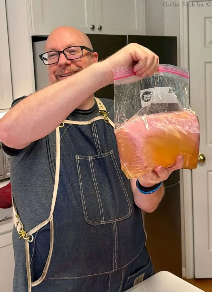 Smiling man in a denim apron holds a large zip-top bag of apple juice brine and a pork loin in a kitchen.