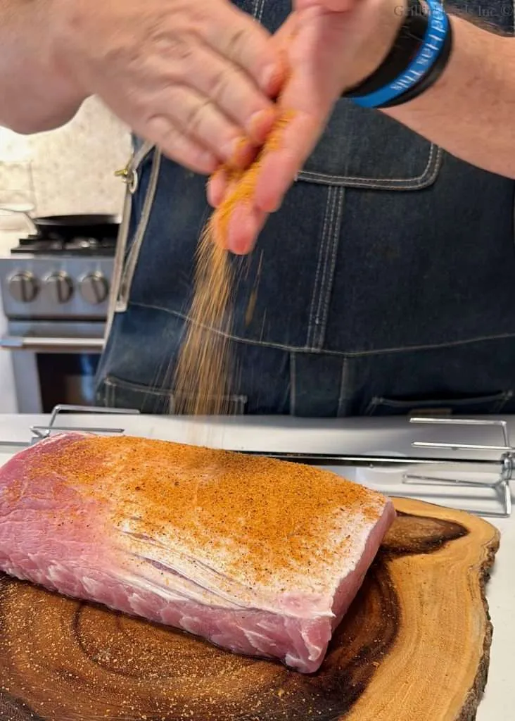 Person sprinkling BBQ seasoning onto a thick pork loin on a wooden cutting board in a kitchen, with a stove in the background.