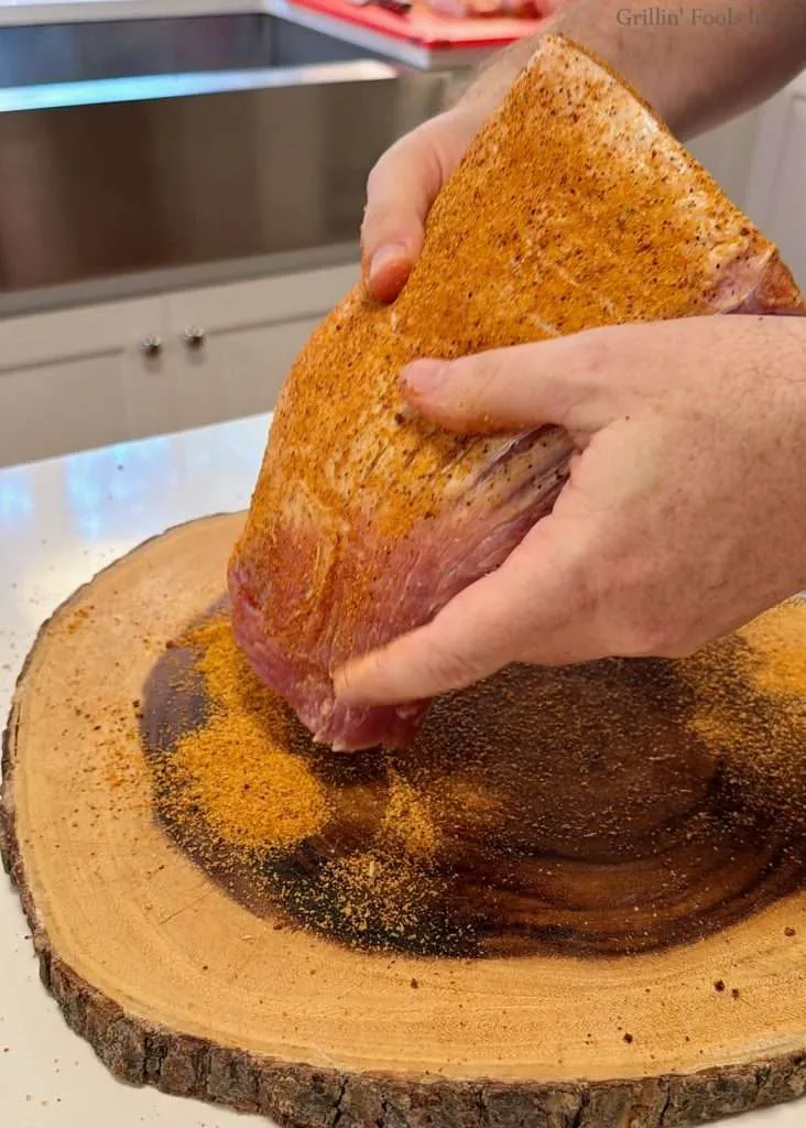 Hands firmly hold a seasoned raw pork loin covered with spices over a round wooden cutting board
