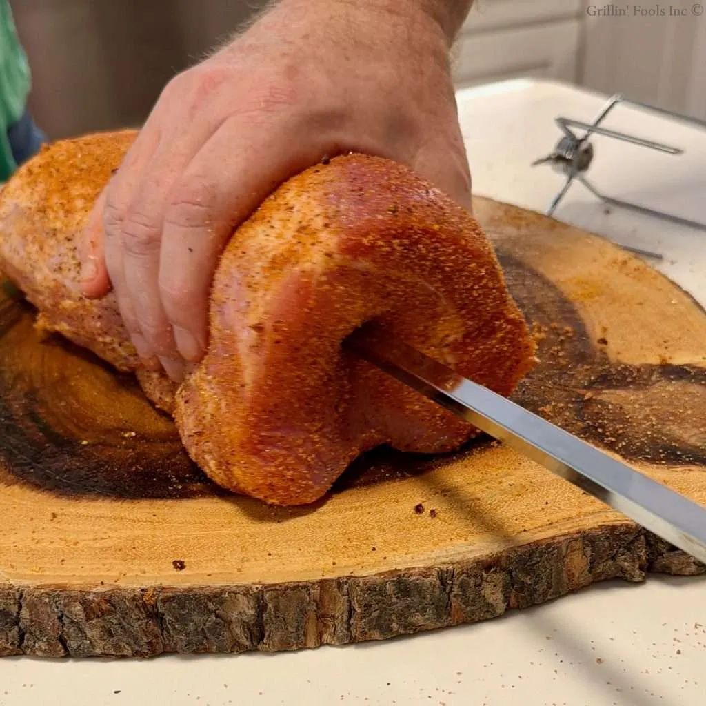 Hand pressing rotisserie spit rod into a heavily seasoned pork loin on a wooden cutting board with a knife nearby in a kitchen setting.